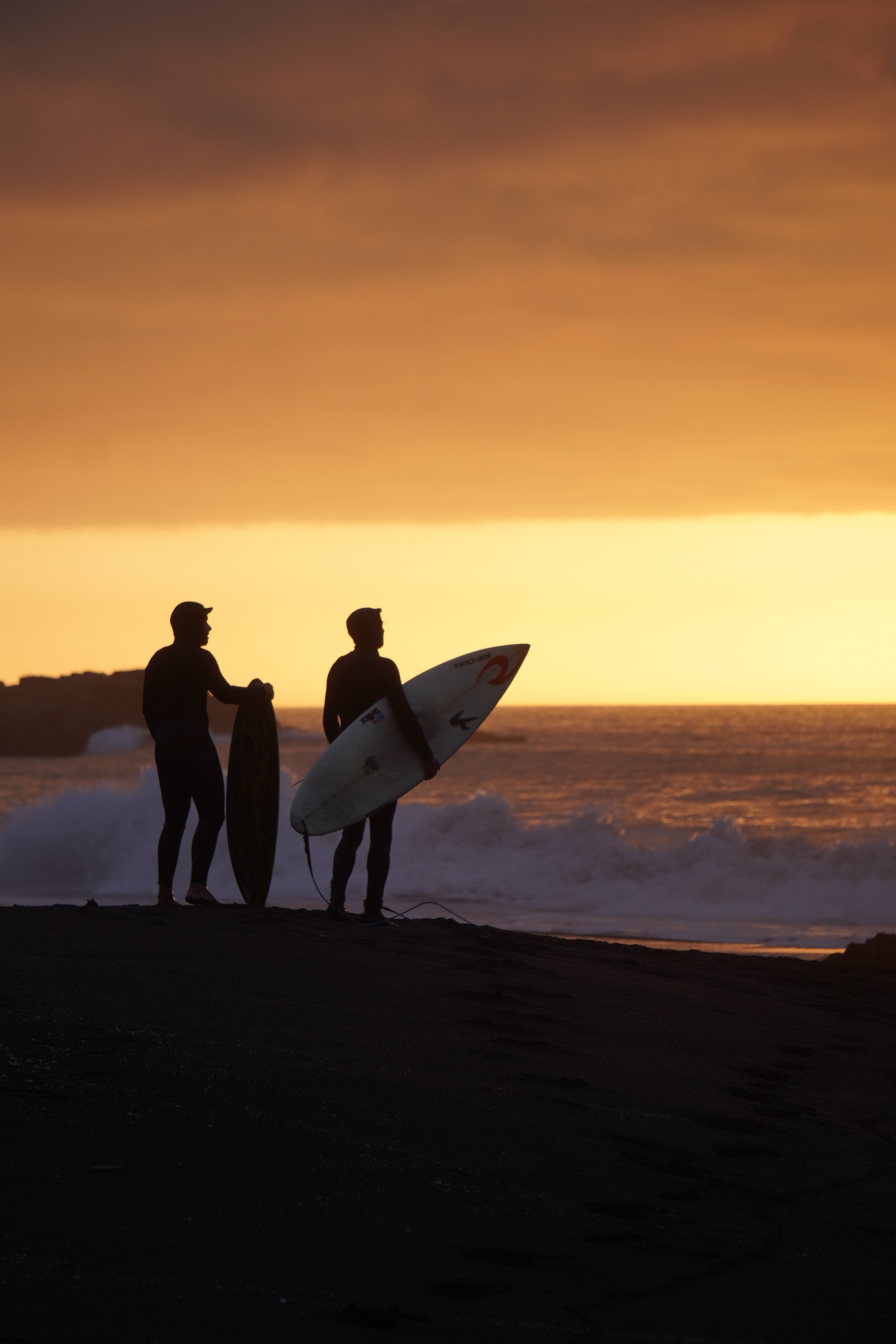 Two surfers on the sand at sunset, boards under their arms