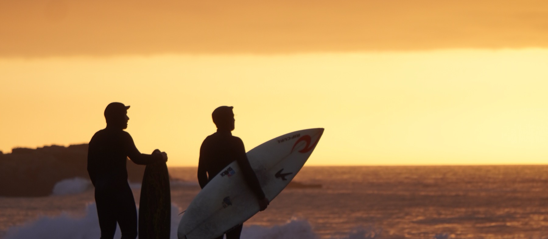 Two surfers on the shore at sunset, boards under their arms