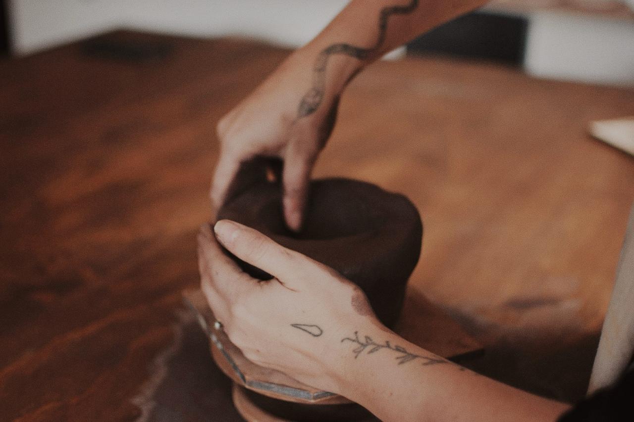 Two hands shaping a dark clay vessel on a wooden table
